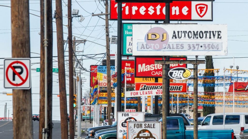 Uma rua movimentada, repleta de cartazes coloridos anunciando empresas automotivas e vendas de carros. No primeiro plano, uma placa de proibido estacionar em um poste.