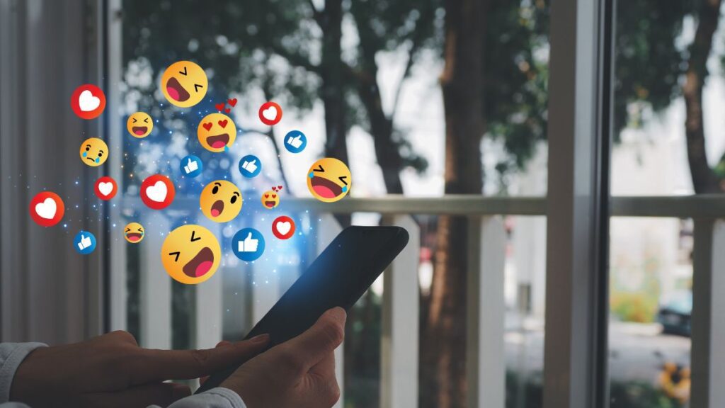 Pessoa usando um smartphone com vários emojis de reação em mídias sociais flutuando acima da tela, indicando interação on-line para promover pequenas empresas.