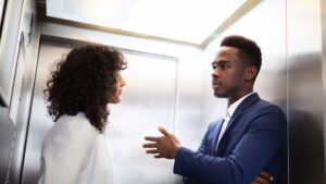 Duas pessoas em trajes de negócios estão conversando dentro de um elevador. A pessoa à direita gesticula com uma mão.